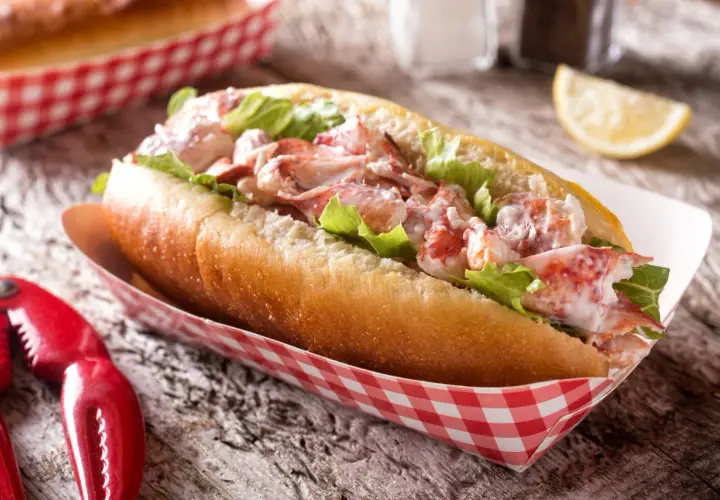 Pyszny lobster roll z kawałkami homara i sałatą w bułce, podany w czerwono-białym papierowym opakowaniu. Obok szczypce do homara.