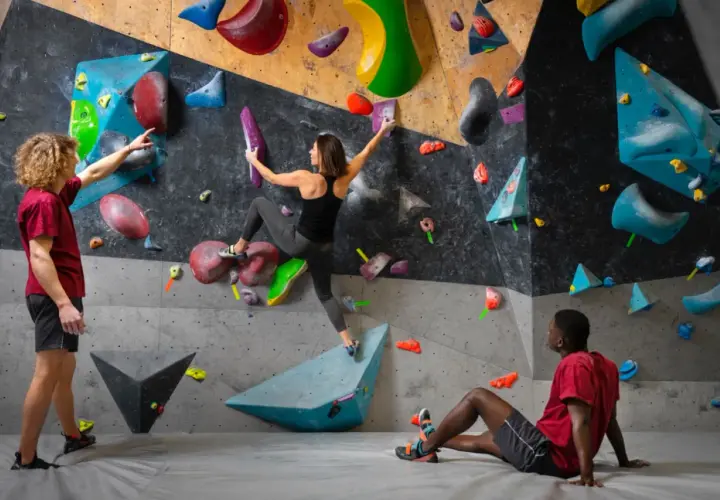 Drei Personen bouldern an einer bunten Wand. Ein Mann gibt Tipps, eine Frau klettert, ein Mann ruht.