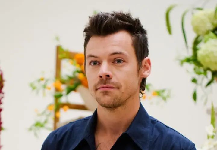 Harry Styles con un moderno mini mullet, mirada intensa y camisa azul, rodeado de arreglos florales.