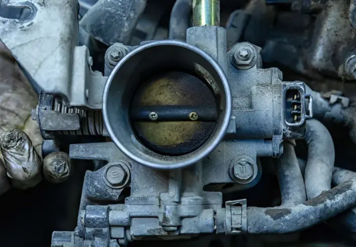 Primer plano de una mariposa de admisión de motor de coche, con suciedad y grasa. Unas manos enguantadas se ven a la izquierda.
