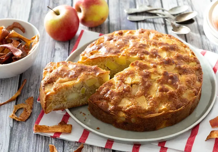 Un délicieux gâteau aux pommes, doré et moelleux, avec des morceaux de fruits visibles. Des pommes fraîches et des chips de pommes complètent la scène.