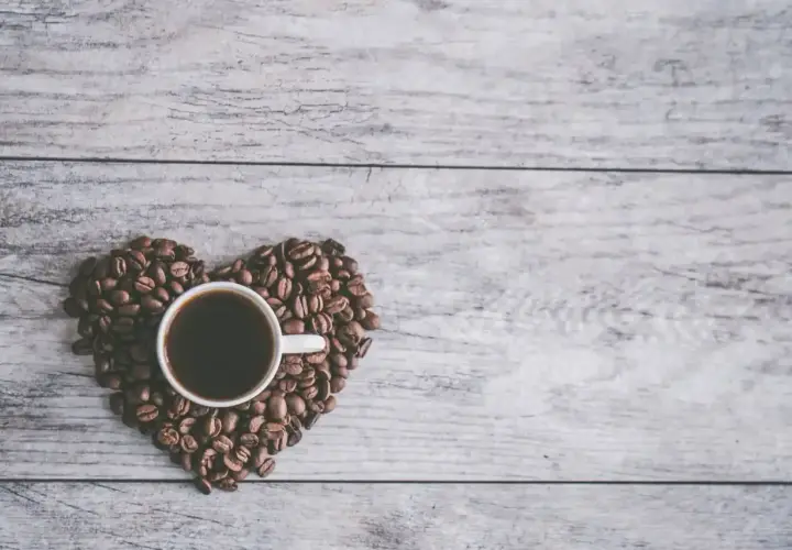 Una tazzina di caffè nero, circondata da chicchi di caffè disposti a forma di cuore su un tavolo di legno. Un invito al caffè in allattamento.