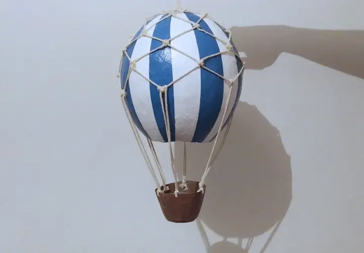 Globo aerostático manualidad, con rayas azules y blancas, suspendido con cuerdas y una cesta de mimbre.