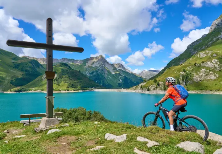 Ciclista en el Alpe Adria Radweg, junto a un lago turquesa y montañas verdes.