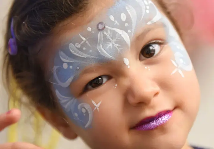 Bambina con trucco da fata del ghiaccio per il carnevale, con glitter viola sulle labbra e disegni bianchi e azzurri sul viso.