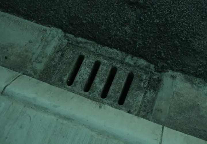 Betonierter Abfluss mit Schlitzen, daneben Asphalt. Der Abfluss erinnert an einen stinkenden revisionsschacht im keller.