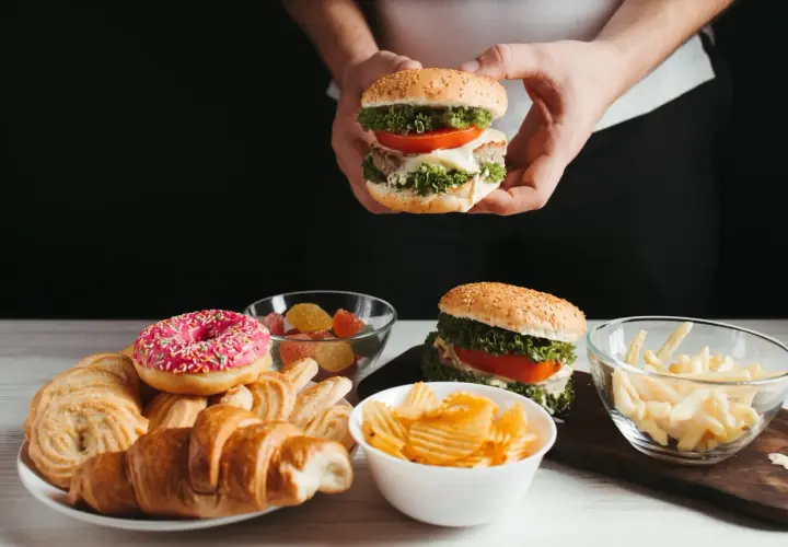 Ręce trzymają burgera, obok talerz z croissantami, pączkiem i ciastkami, miseczki z żelkami, frytkami i chipsami. Czy cheat day przyspiesza metabolizm?
