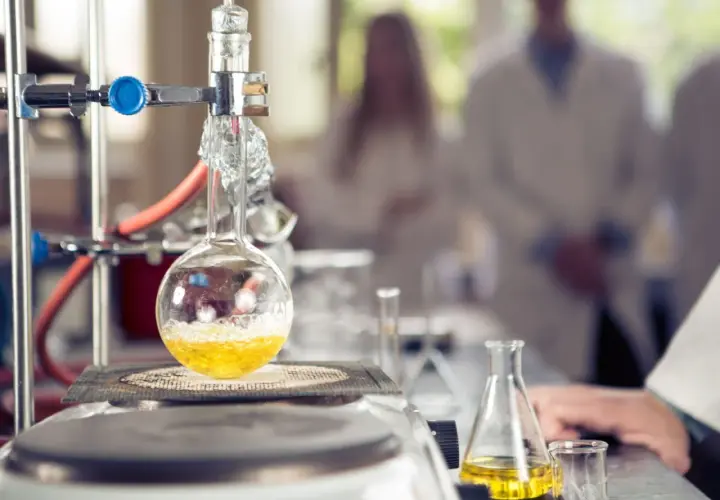 W laboratorium trwa eksperyment. W kolbie okrągłodennej, podgrzewanej na płycie, bulgocze żółty płyn. W tle widać naukowców w kitlach, badających wartościowość pierwiastków.