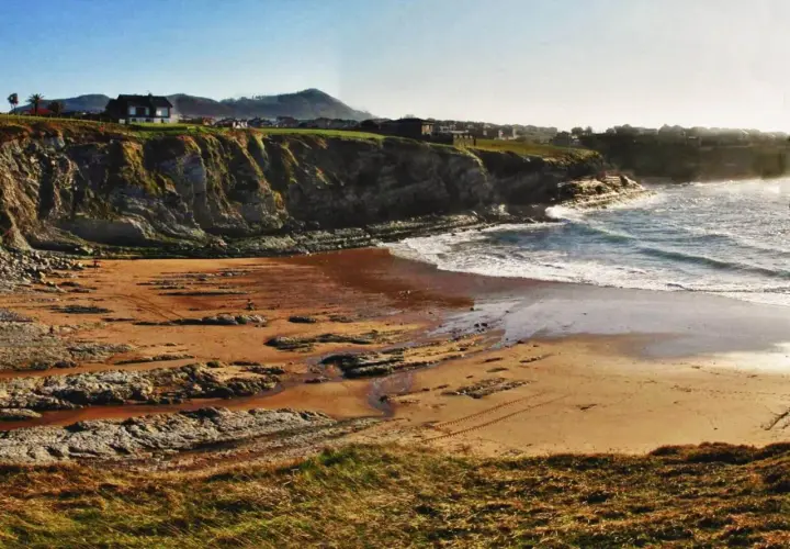 La playa de Portio, con sus acantilados rocosos y arena dorada, se baña en la luz del sol. Las olas rompen suavemente en la costa.