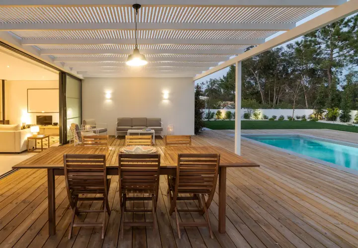 Table et chaises en bois huilé sur une terrasse en bois, avec piscine et jardin en arrière-plan.