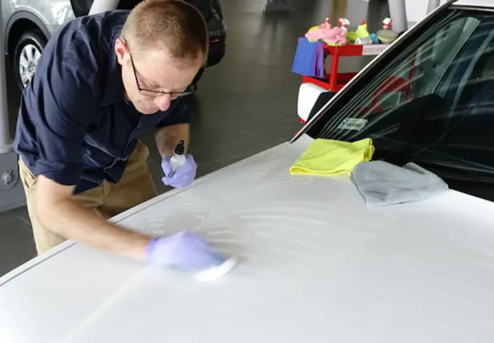 Mężczyzna w rękawiczkach nakłada powłokę ceramiczną na maskę białego samochodu, używając aplikatora i buteleczki z płynem.