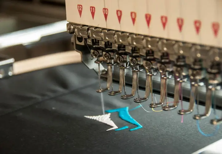 Machine à broder en action, créant un motif coloré sur un tissu noir. La **direct to garment printing** permet des personnalisations uniques.