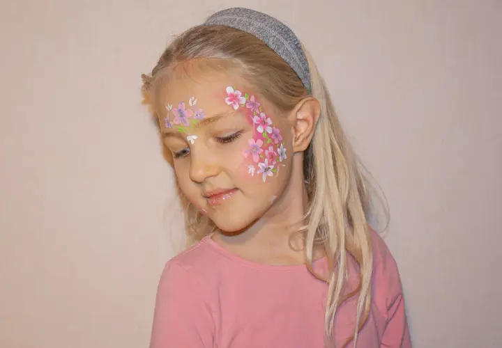 Ein Mädchen mit blonden Haaren und einem Stirnband, das ein Blumenmuster auf der Wange trägt. Das Stern schminken Kinder ist hier als zarte Blüten umgesetzt.
