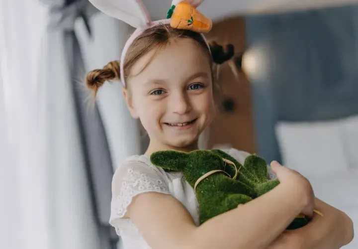 Uśmiechnięta dziewczynka w króliczych uszkach przytula zielonego zajączka. Wesoła wielkanoc w hotelu.