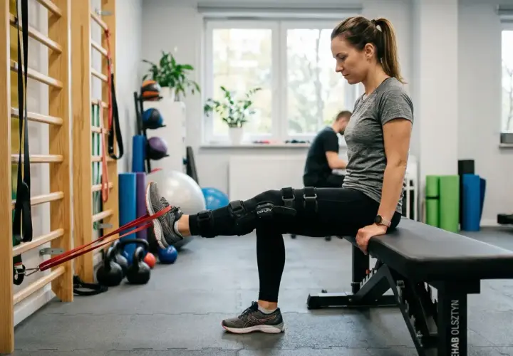 Kobieta w ortezie kolana wykonuje ćwiczenia z taśmą oporową, rehabilitując gonartrozę.