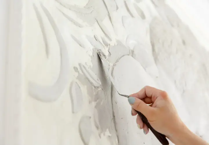 Mano che lavora con uno strumento su uno stucco murale in rilievo, creando un'opera d'arte tridimensionale.