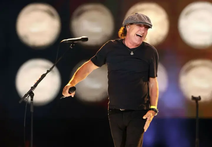 Brian Johnson w czapce i czarnej koszulce śpiewa do mikrofonu na scenie, rozbawiony.
