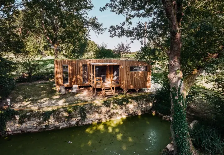Une maison bioclimatique en bois, perchée près d'un étang verdoyant, entourée d'arbres majestueux.