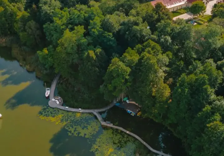 Widok z lotu ptaka na jezioro z żaglówką, drewnianą kładką i otaczającym lasem.