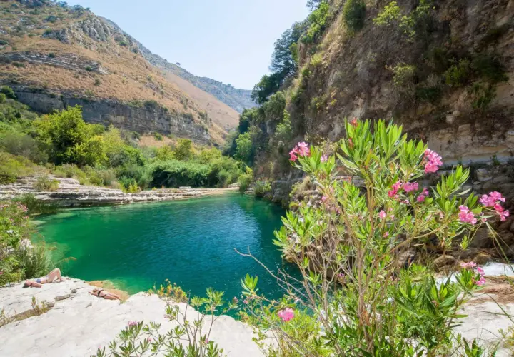 A serene pool of turquoise water reflects the rugged beauty of Sicily nature. Two sunbathers relax on the rocky shore, surrounded by lush greenery and blooming pink oleander flowers.