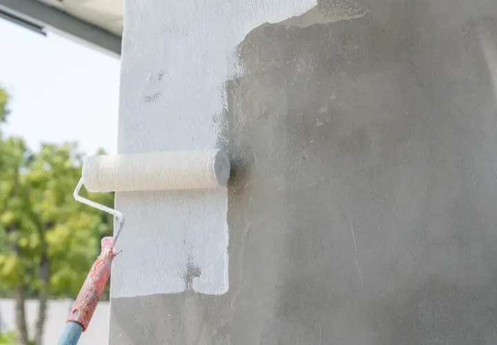 Wałek malarski pokrywa białą farbą szary beton. Prace wykończeniowe na zewnątrz budynku.