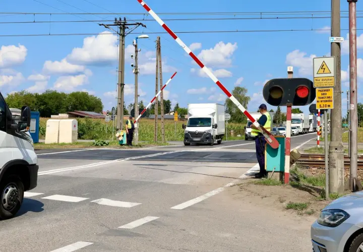 Wjazd na przejazd kolejowy. Zapory opuszczone, czerwone światło sygnalizatora. Pracownik w kamizelce odblaskowej pilnuje ruchu.