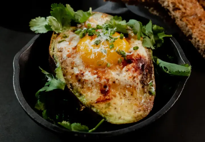 Un avocat oeuf au four, garni de coriandre fraîche et de ciboule, servi dans une poêle en fonte avec du pain grillé.