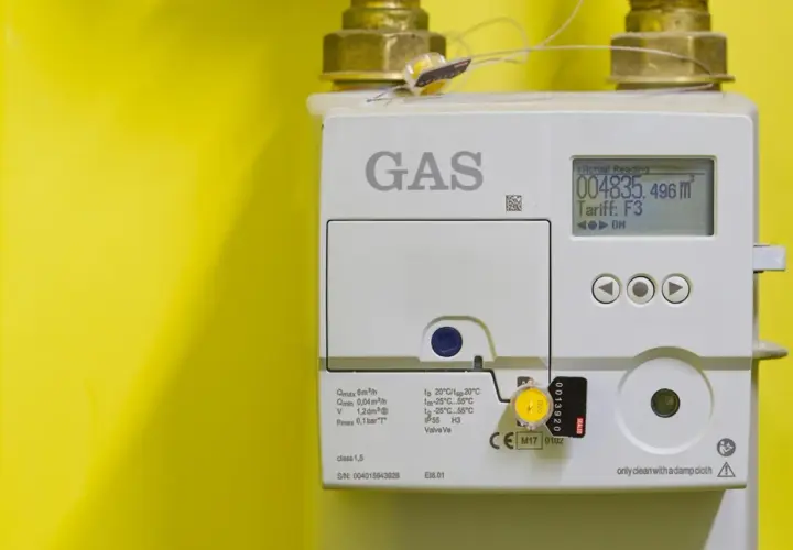Medidor de gas blanco con pantalla digital y conexiones doradas sobre fondo amarillo.