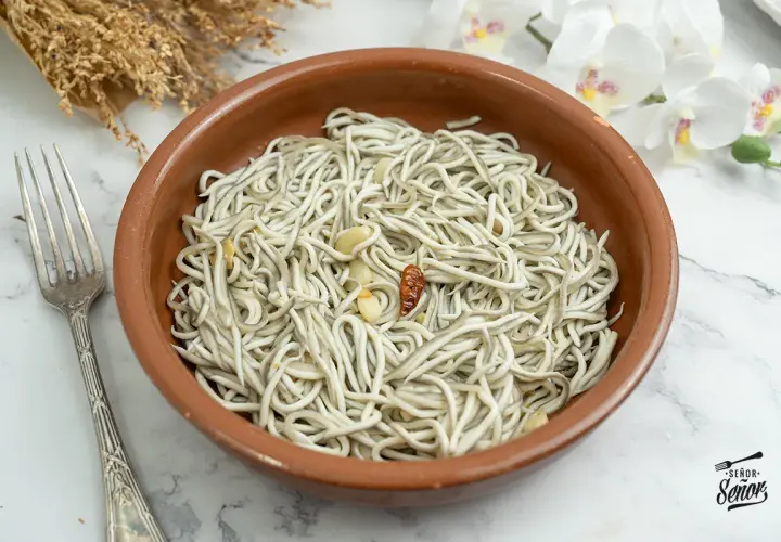 Un cuenco de barro con gulas al ajillo, ajo y guindilla. Un tenedor plateado a la izquierda y flores blancas al fondo.