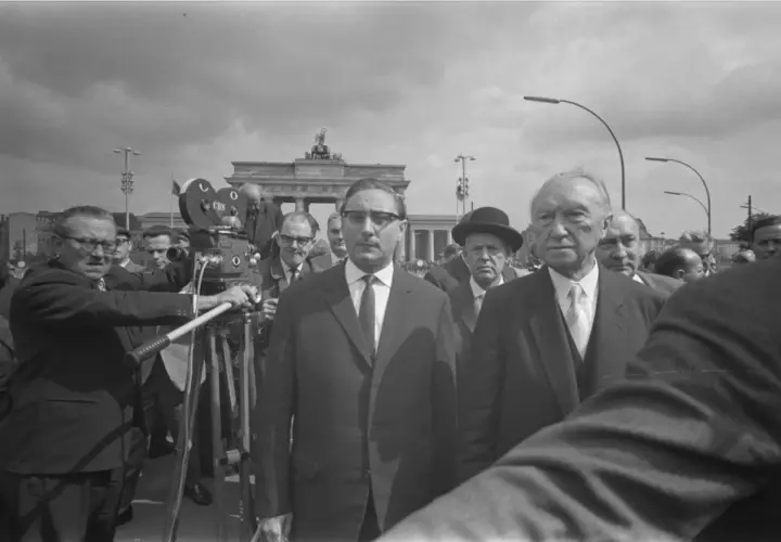 Männer in Anzügen, eine Filmkamera und das Brandenburger Tor im Hintergrund. Ein Mann hält eine "Stalin-Note" in der Hand.
