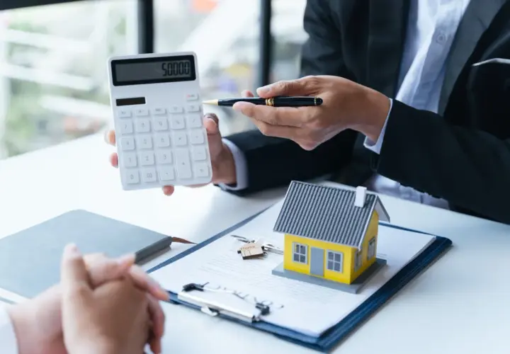 Conseiller montrant un calcul sur une calculatrice pour une aide premier achat immobilier. Une maison miniature et des clés sont sur un contrat.