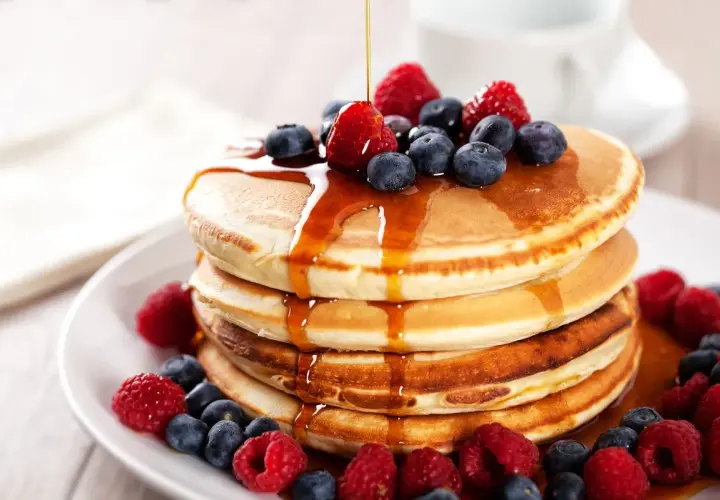 Pancake ricetta: una pila di soffici pancake guarniti con sciroppo d'acero, lamponi e mirtilli freschi.