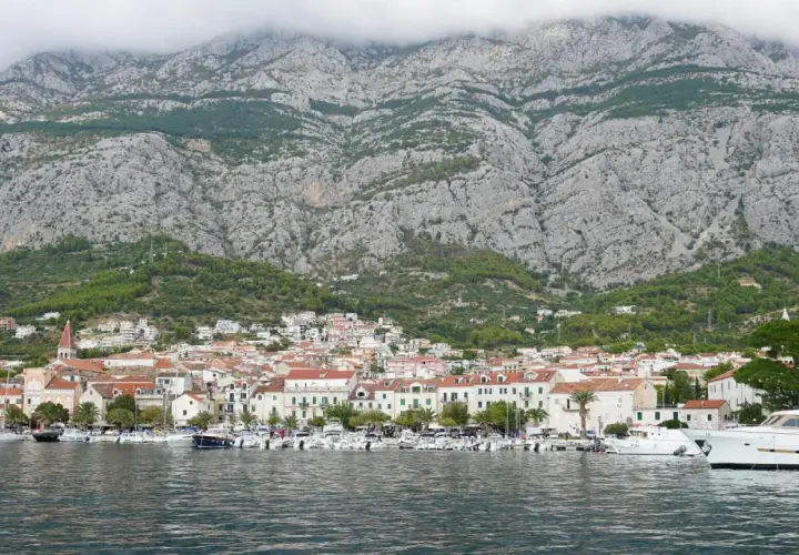 Widok na miasto Makarska z górami w tle. W porcie cumują liczne łodzie i jachty. Choć nie ma tu lotniska, widok zapiera dech w piersiach.