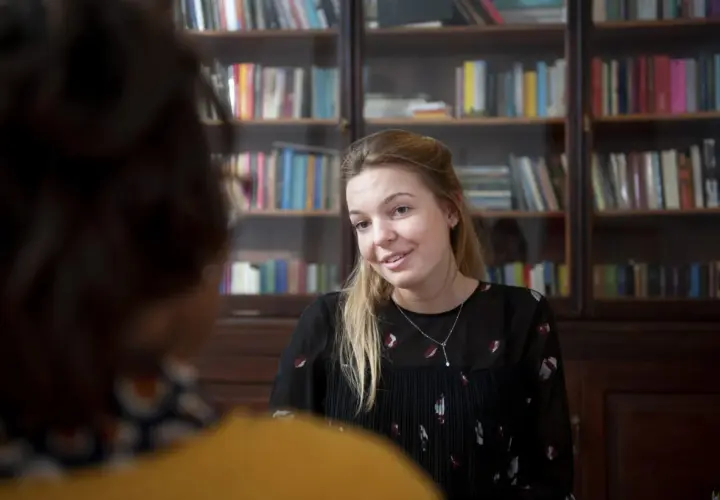 Kobieta z blond włosami, w czarnej bluzce, rozmawia z kimś, kto siedzi tyłem. Za nią regały z książkami. Może to być przykład odwróconej psychologii w związku.