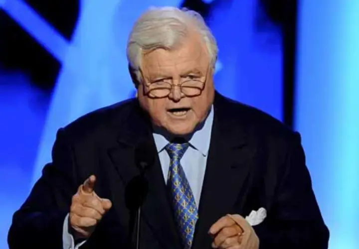 Ein älterer Mann mit Brille und grauem Haar spricht in ein Mikrofon. Er trägt einen dunklen Anzug mit einer blauen Krawatte. Seine Hand ist erhoben, als würde er eine Rede halten, die an die Kennedy-Krankheit erinnert.