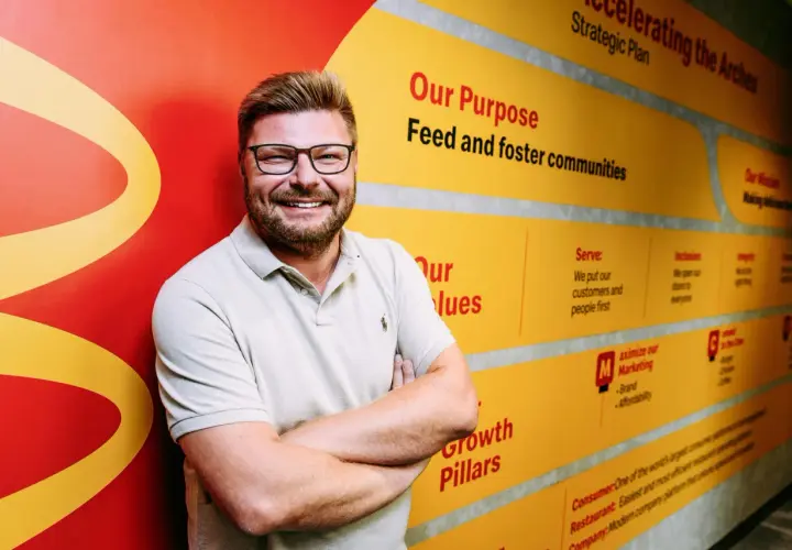 Uśmiechnięty mężczyzna w okularach na tle ściany z hasłami McDonald's. Zastanawiasz się, ile jest McDonaldów w Polsce? Ta ściana może pomóc w rozwoju sieci.