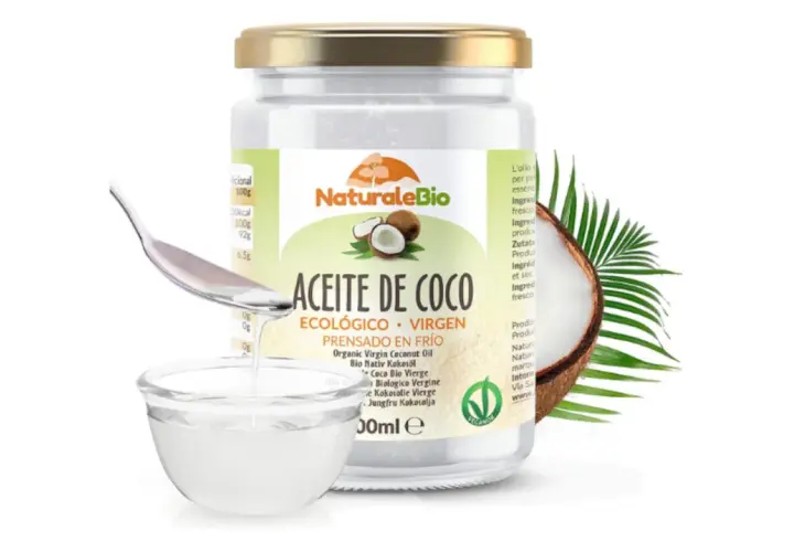 Un frasco de aceite de coco ecológico y virgen, ideal para el cuidado del pelo. Una cuchara vierte el aceite en un cuenco.