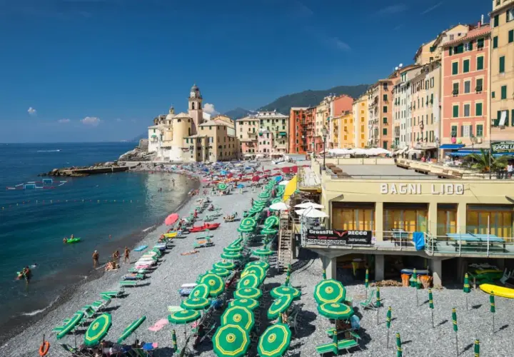 Plage animée à Camogli, Ligurie, avec des parasols verts et jaunes, des bâtiments colorés et la mer bleue.