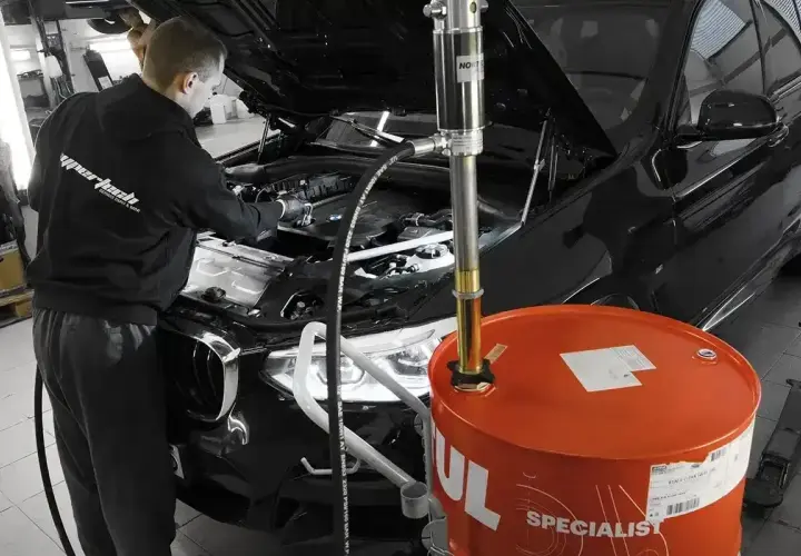 Mechanik wymienia olej w czarnym BMW. Obok stoi duża, pomarańczowa beczka z olejem. Pamiętaj, co ile wymiana oleju jest kluczowa dla silnika.