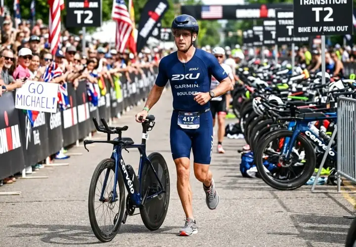 Zawodnik w stroju do triathlonu olimpijskiego biegnie z rowerem obok rzędu rowerów. Tłum kibicuje, trzymając transparent "GO MICHAEL!".