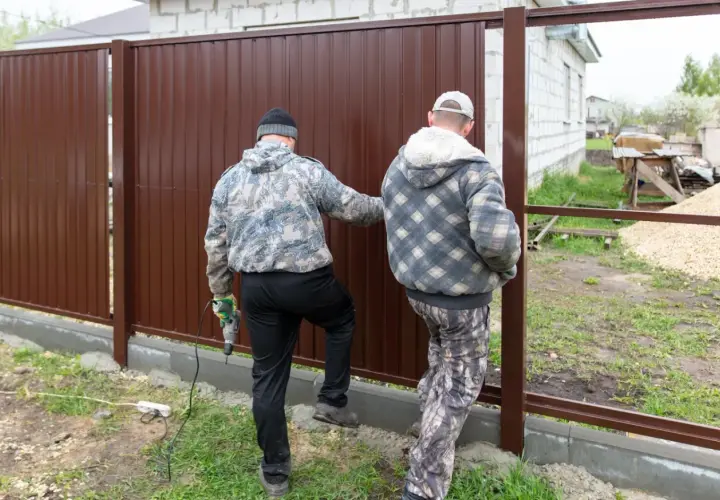 Dwóch mężczyzn montuje brązowe ogrodzenie. Jeden z nich trzyma wiertarkę. Ogrodzenie niezgodne z granicą działki, budowa w toku.