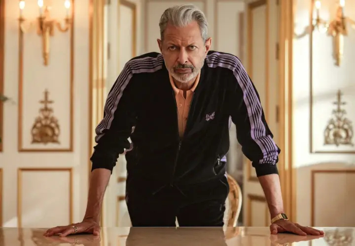 Jeff Goldblum w dresie z fioletowymi pasami, jak z jego filmów, opiera się o stół w eleganckim wnętrzu.