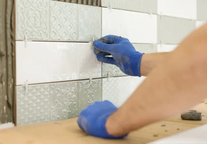 Ręce w niebieskich rękawiczkach wykonują naprawę płytek ceramicznych, precyzyjnie układając nowe elementy na ścianie.