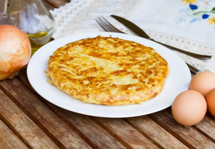 Frittata di patate dorata su piatto bianco, con cipolla, uova e olio d'oliva come ingredienti.