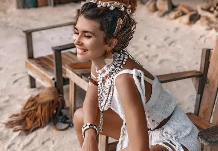 Uśmiechnięta kobieta w stylu boho, ozdobiona muszlami i piórami, siedzi na plaży.