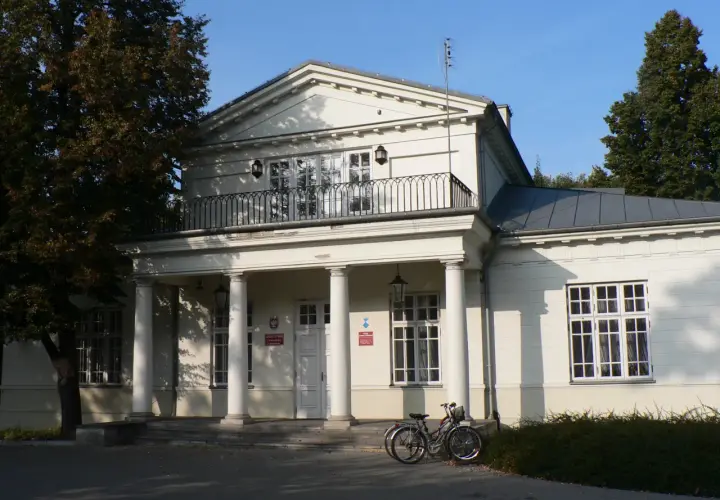 Pałac Potulickich w Pruszkowie, z kolumnowym portykiem i balkonem, otoczony zielenią. Dwa rowery stoją przed wejściem.
