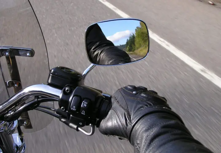 En la carretera, un motorista con guantes de cuero revisa el único espejo de su motocicleta, reflejando el cielo y los árboles.