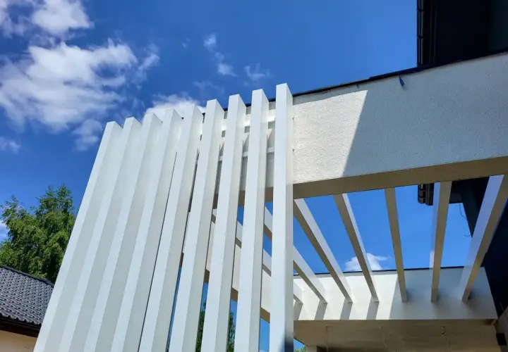 Nowoczesna pergola z białymi, pionowymi lamelami, wykonana z **belki aluminiowe**. Konstrukcja tworzy ciekawy wzór na tle błękitnego nieba z chmurami.