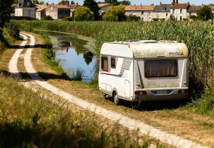 Caravane stationnée au bord d'un canal, dans le Marais Poitevin. Un chemin de terre serpente à travers l'herbe haute.