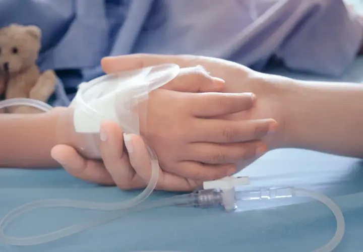 Dłoń dorosłego obejmuje dłoń dziecka z wenflonem. W tle pluszowy miś. To obraz wsparcia, gdy potrzebny jest zasilek pielegnacyjny.